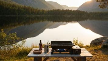 Kompakt bærbar gassgrill på et trebord ved en norsk fjellsjø i gylden kveldssol